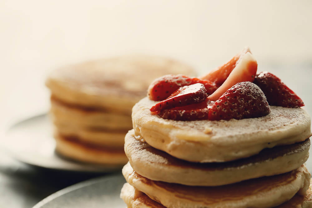 A stack of fluffy pancakes topped with fresh, sliced strawberries and a light dusting of powdered sugar. The pancakes appear golden-brown and are set against a softly blurred background featuring another stack of pancakes, giving a warm and inviting feel to the image.