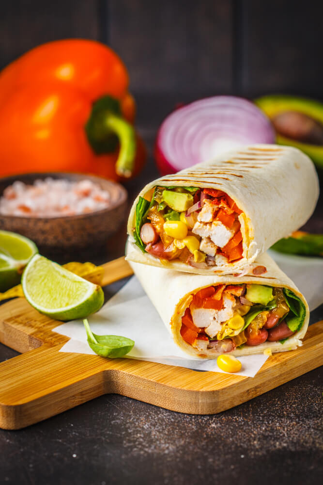 Breakfast burrito filled with grilled chicken, avocado, corn, beans, and vegetables, served on a wooden cutting board with lime wedges.