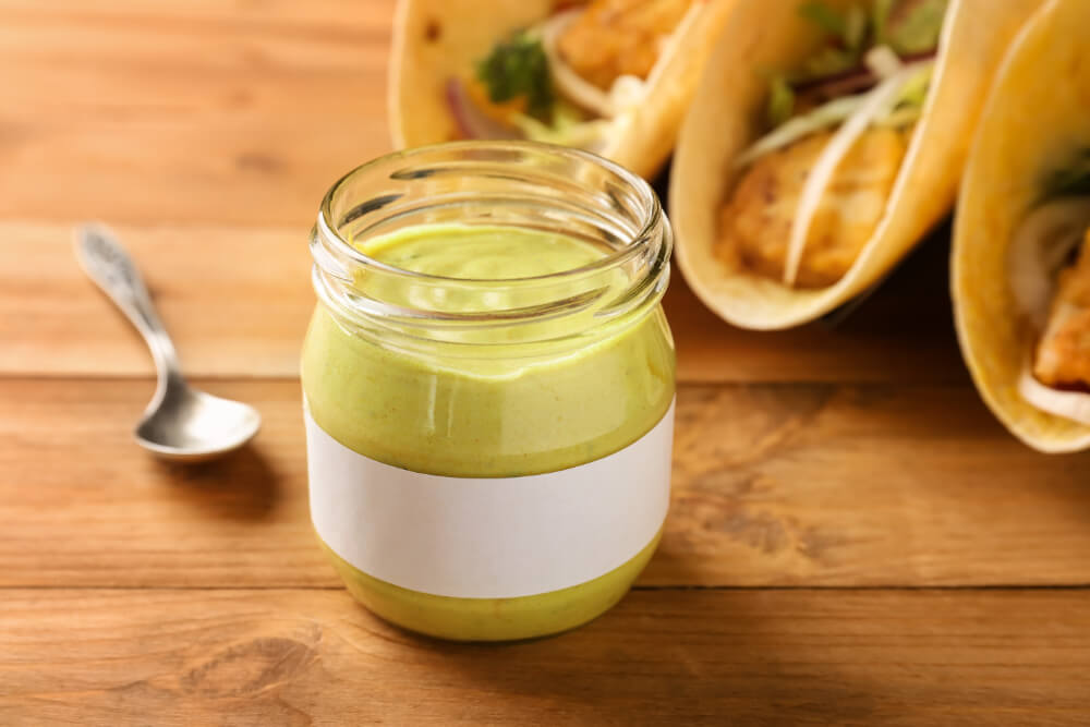 A jar of creamy green taco sauce with a blank label in the foreground, surrounded by fresh fish tacos filled with crispy fish, vegetables, and tortillas on a wooden surface.