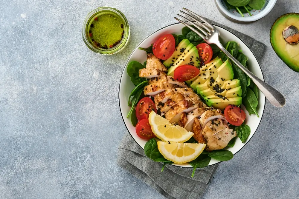 A healthy protein salad with grilled chicken breast, avocado slices, cherry tomatoes, spinach leaves, black sesame seeds, and lemon wedges on a gray background.