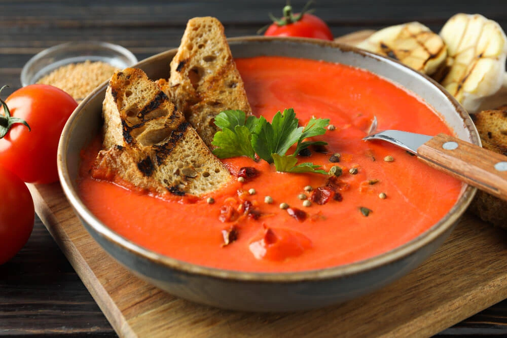 A vibrant bowl of tomato bisque soup garnished with parsley, grilled bread slices, and spices, surrounded by fresh tomatoes and garlic