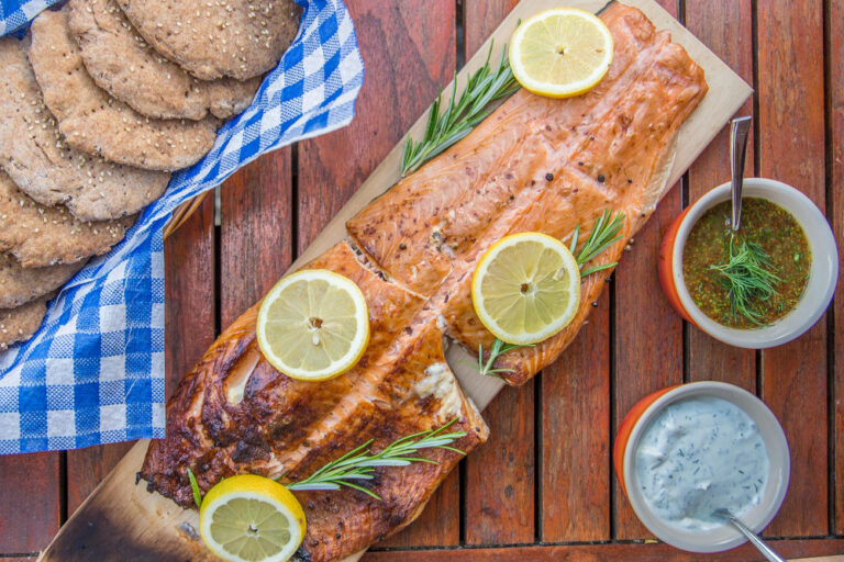 Grilled salmon fillet garnished with lemon slices and rosemary, served with sauces and fresh bread on a wooden table.