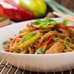 A bowl of stir-fried noodles with chicken, colorful vegetables, and garnished with fresh green onions. Chow Mein vs Lo Mein