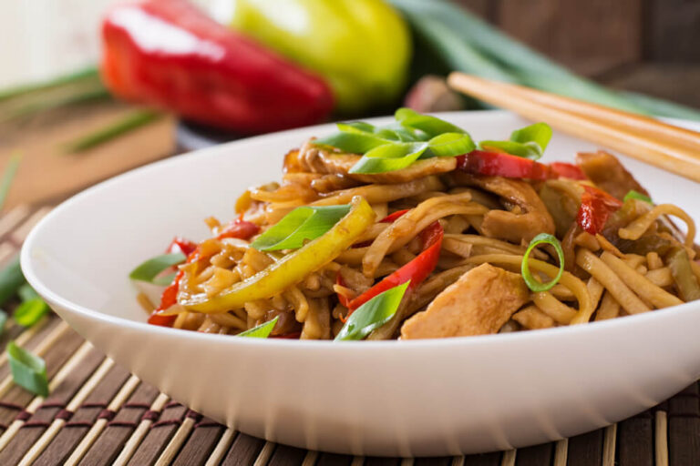 A bowl of stir-fried noodles with chicken, colorful vegetables, and garnished with fresh green onions. Chow Mein vs Lo Mein