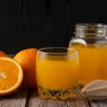 A glass of fresh orange juice with whole oranges and a wooden juicer on a rustic wooden table.