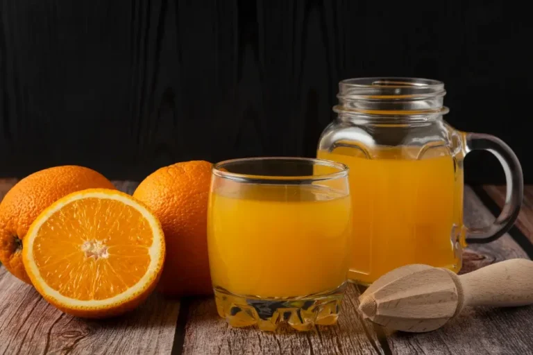 A glass of fresh orange juice with whole oranges and a wooden juicer on a rustic wooden table.