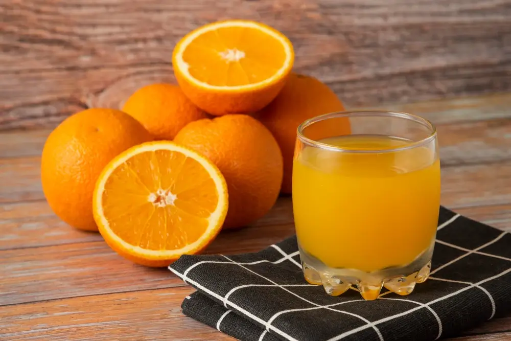 A glass of fresh orange juice placed on a checkered napkin with whole and halved oranges in the background