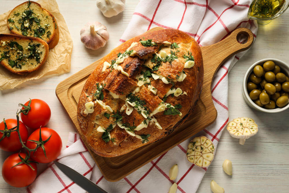 Homemade pull apart garlic bread stuffed with butter, garlic, and herbs, served on a wooden board with fresh tomatoes, garlic bulbs, and olives.