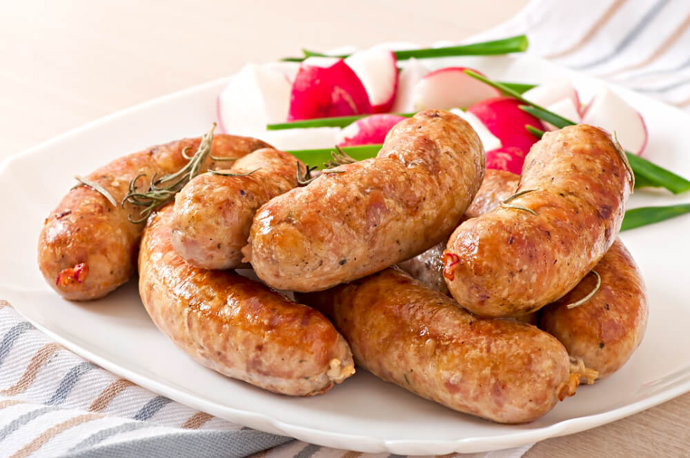 Grilled pork sausages served on a white plate with fresh radishes, green onions, and garnished with rosemary sprigs.