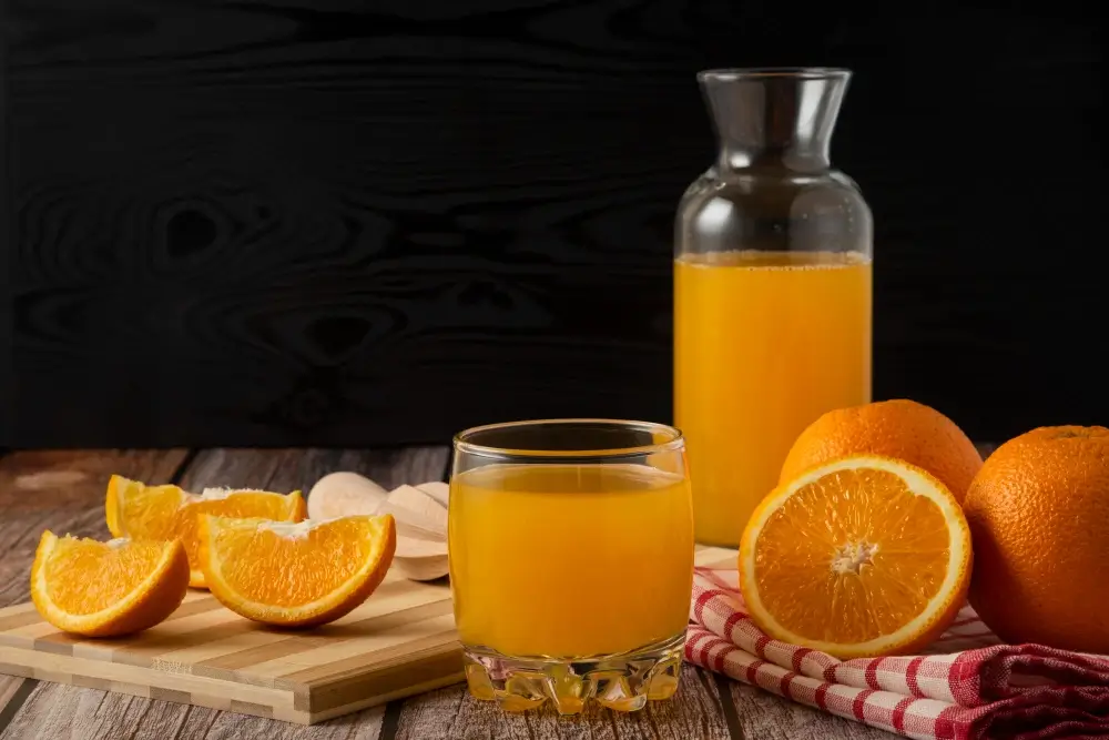 Fresh orange juice in a glass and a carafe with orange slices and whole oranges on a rustic wooden table."