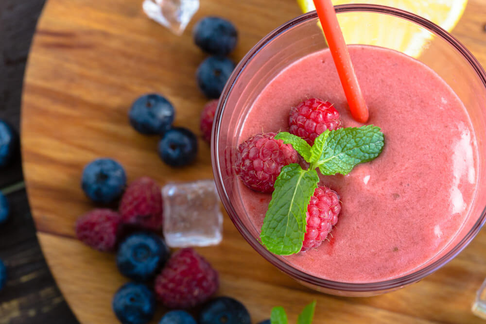 A vibrant pink berry smoothie topped with fresh raspberries, mint leaves, and an orange straw, surrounded by blueberries and ice cubes on a wooden surface.