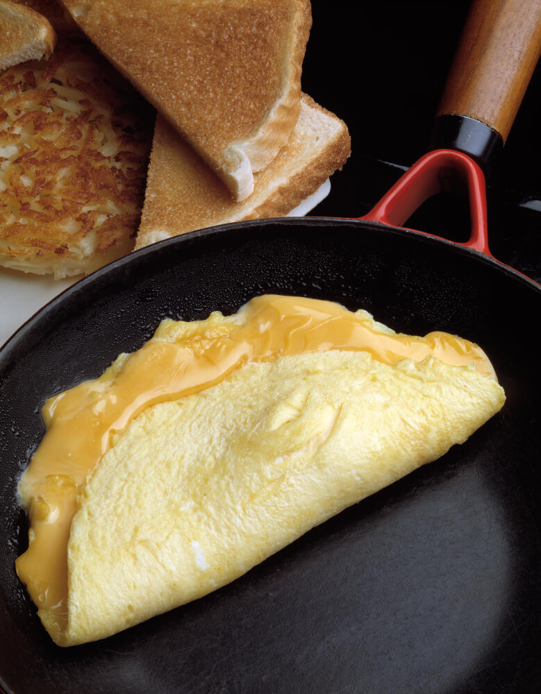 A golden cheese omelette cooking in a skillet, with melted cheese oozing out, served alongside toasted bread and crispy hash browns.