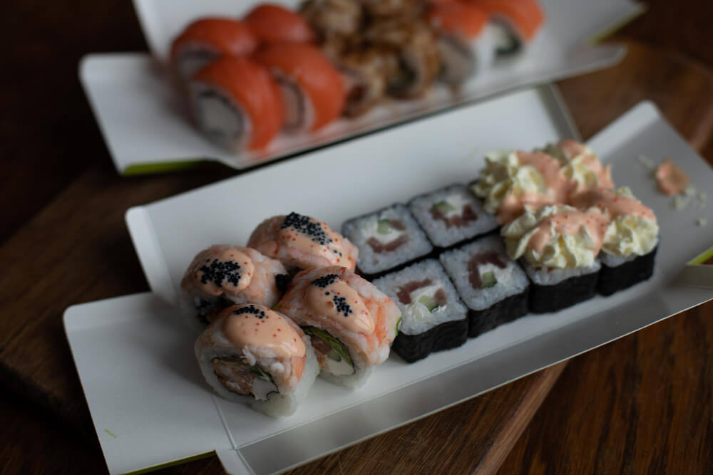 Assorted cooked sushi rolls with creamy toppings, salmon, and seaweed-wrapped pieces served in a takeout container.