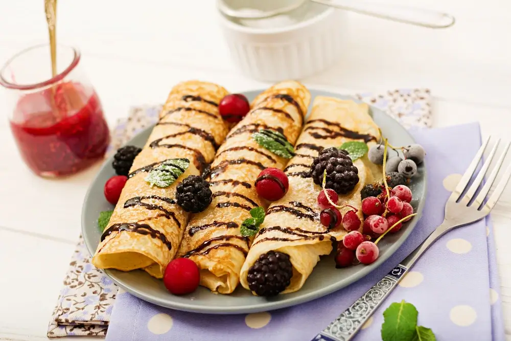 Rolled Swedish pancakes on a plate, topped with chocolate drizzle, blackberries, red currants, blueberries, and mint leaves, with a jar of jam in the background."