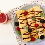 "Top view of rolled Swedish pancakes drizzled with chocolate and garnished with blackberries, red currants, blueberries, and mint leaves, accompanied by a jar of jam."