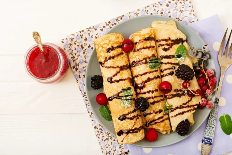 "Top view of rolled Swedish pancakes drizzled with chocolate and garnished with blackberries, red currants, blueberries, and mint leaves, accompanied by a jar of jam."