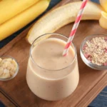 "A glass of creamy banana oatmeal smoothie with a striped straw, surrounded by bananas, sliced almonds, and a bowl of oats with berries, placed on a wooden tray."