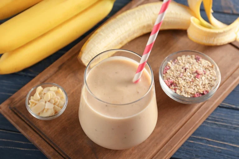 "A glass of creamy banana oatmeal smoothie with a striped straw, surrounded by bananas, sliced almonds, and a bowl of oats with berries, placed on a wooden tray."