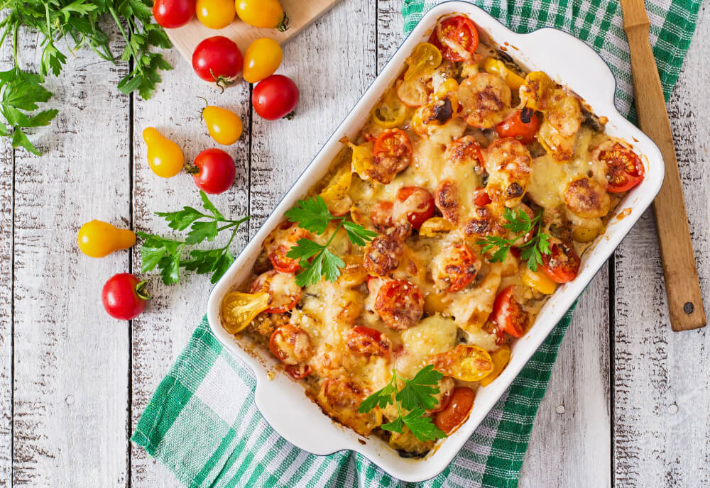 A vibrant baked casserole with cherry tomatoes, sausages, melted cheese, and fresh parsley in a white baking dish on a green checkered cloth.