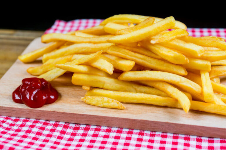 A pile of golden French fries served on a wooden board with a dollop of ketchup, placed on a red-and-white checkered tablecloth.