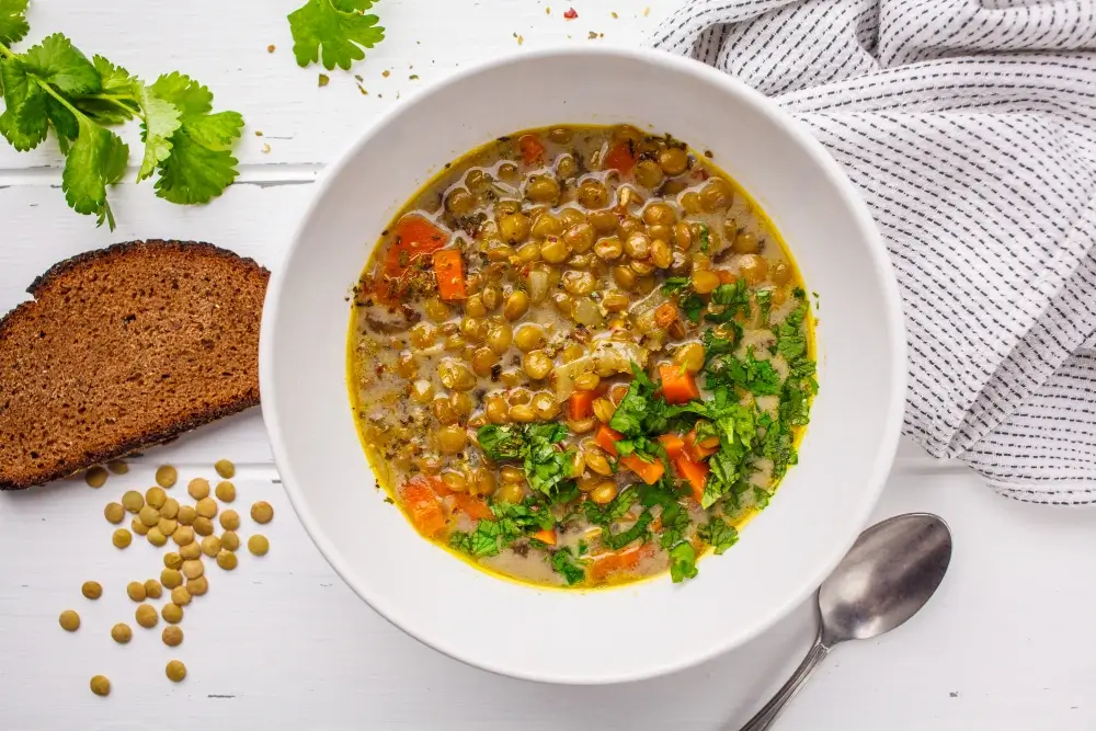 A bowl of lentil vegetable soup garnished with fresh cilantro, served with a slice of rye bread and scattered lentils on a white background.