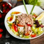 A protein salad with tuna, hard-boiled eggs, cherry tomatoes, cucumber, lettuce, and a lemon wedge, served on a wooden table with scattered cherry tomatoes in the background.