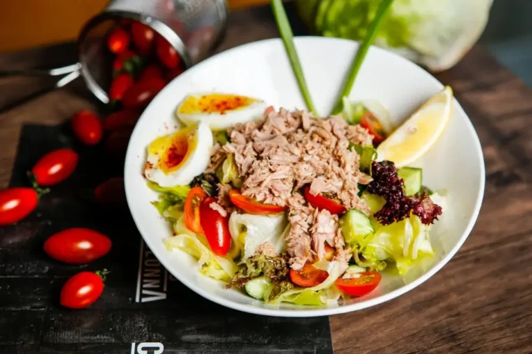 A protein salad with tuna, hard-boiled eggs, cherry tomatoes, cucumber, lettuce, and a lemon wedge, served on a wooden table with scattered cherry tomatoes in the background.