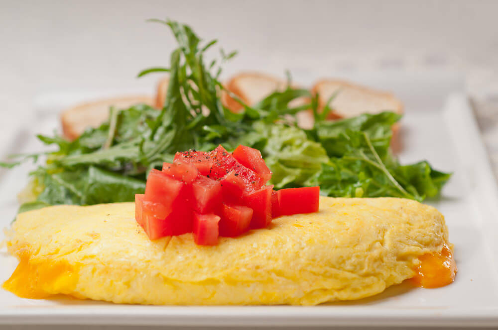 A fluffy cheese omelette served with diced tomatoes on top, accompanied by a fresh arugula salad and slices of toasted bread on a white plate.