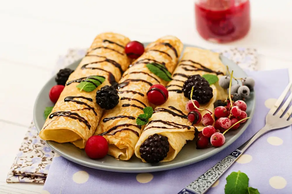 Swedish pancakes rolled and topped with chocolate drizzle, blackberries, red currants, blueberries, and mint leaves on a decorative plate."