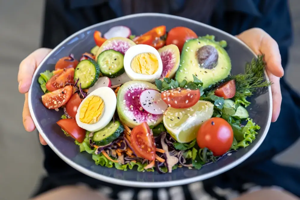 A vibrant protein salad with hard-boiled eggs, avocado, radishes, cucumbers, cherry tomatoes, and fresh greens, garnished with sesame seeds and served with lemon wedges.