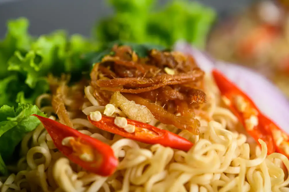 A close-up of Chicken Lo Mein topped with shredded chicken, red chili slices, and fresh lettuce.