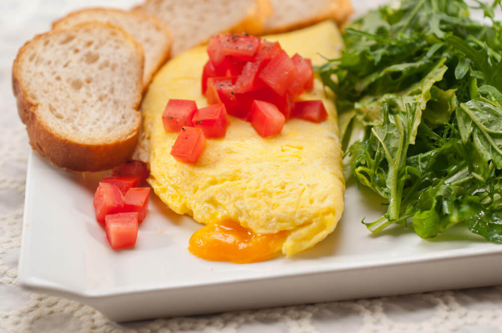 A golden cheese omelette topped with fresh diced tomatoes, served with a side of arugula salad and toasted bread slices on a white plate.