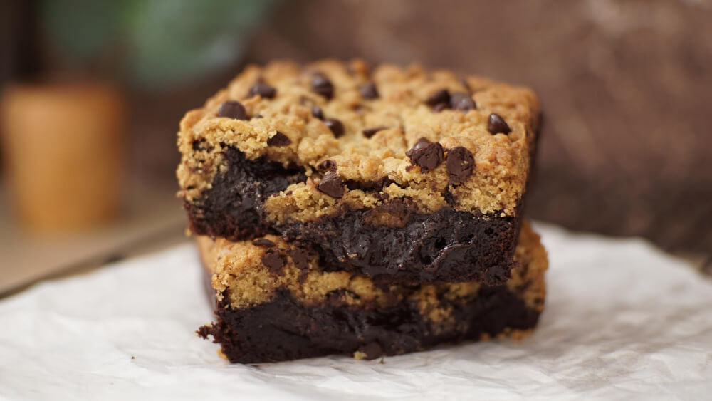 Two chocolate chip cookie brownies stacked on parchment paper, showcasing a gooey chocolate center and golden cookie layer.