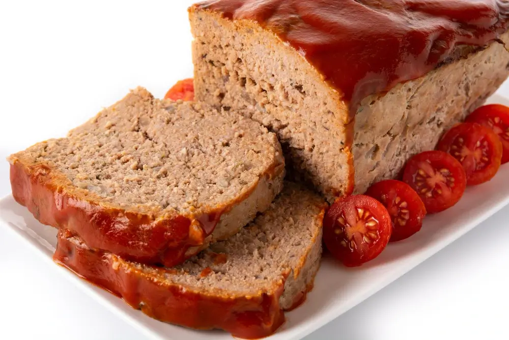 Close-up of a meatloaf topped with a shiny tomato glaze, sliced and served with cherry tomatoes on a white plate.