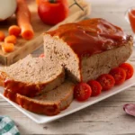 A freshly baked meatloaf topped with a glossy red glaze, sliced and served on a white plate, garnished with cherry tomatoes. Ingredients like carrots, onions, and garlic are displayed in the background.