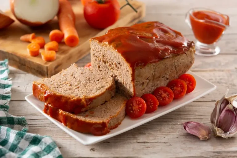 A freshly baked meatloaf topped with a glossy red glaze, sliced and served on a white plate, garnished with cherry tomatoes. Ingredients like carrots, onions, and garlic are displayed in the background.