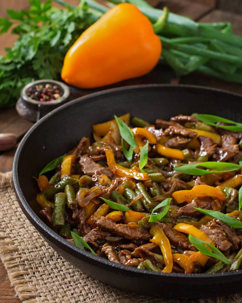 A close-up of a vibrant beef stir fry in a black skillet, featuring tender beef strips, green beans, yellow bell peppers, and garnished with fresh herbs.