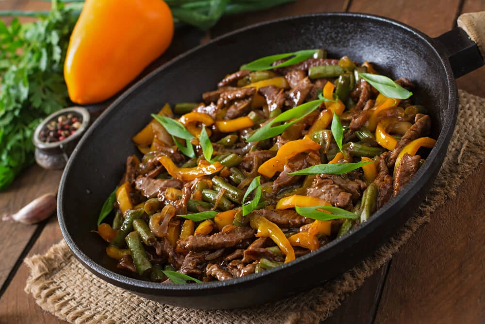 A skillet filled with a vibrant beef stir fry, featuring tender beef slices, crisp green beans, yellow bell peppers, and fresh herb garnishes.