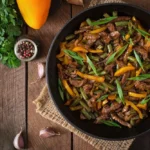 A skillet filled with colorful beef stir fry, including thinly sliced beef, yellow bell peppers, green beans, and garnished with fresh green herbs.