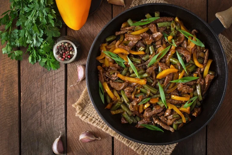 A skillet filled with colorful beef stir fry, including thinly sliced beef, yellow bell peppers, green beans, and garnished with fresh green herbs.