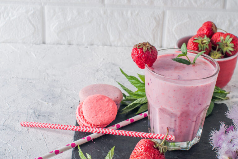 A creamy strawberry smoothie in a glass, garnished with fresh strawberries and mint, with pink macarons and red-and-white striped straws on a slate surface.