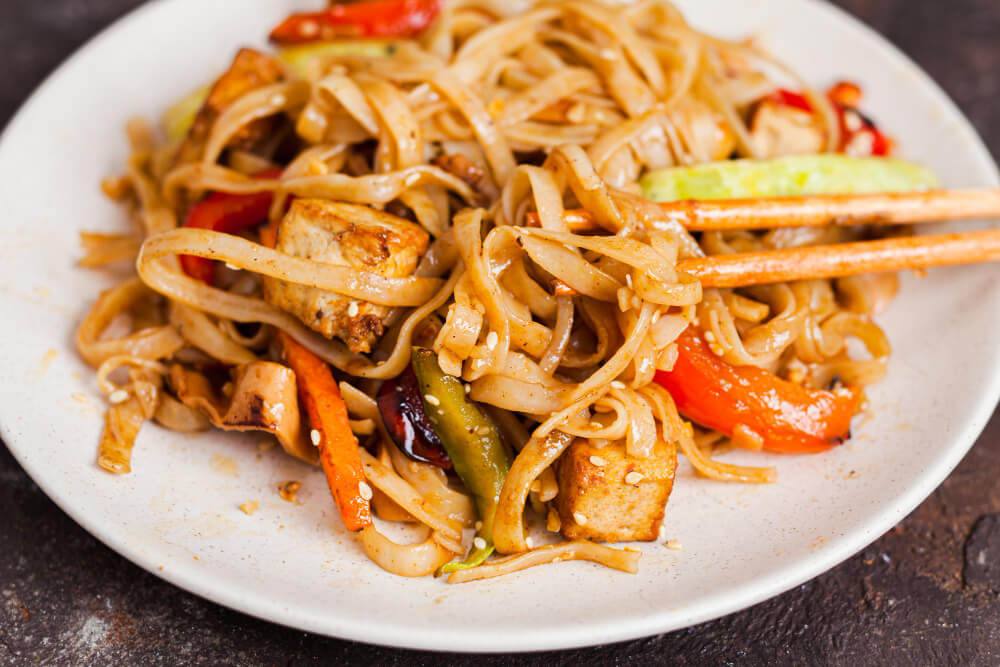 This is a close-up of a plate of stir-fried noodles with vegetables, tofu, and sesame seeds, served with chopsticks.