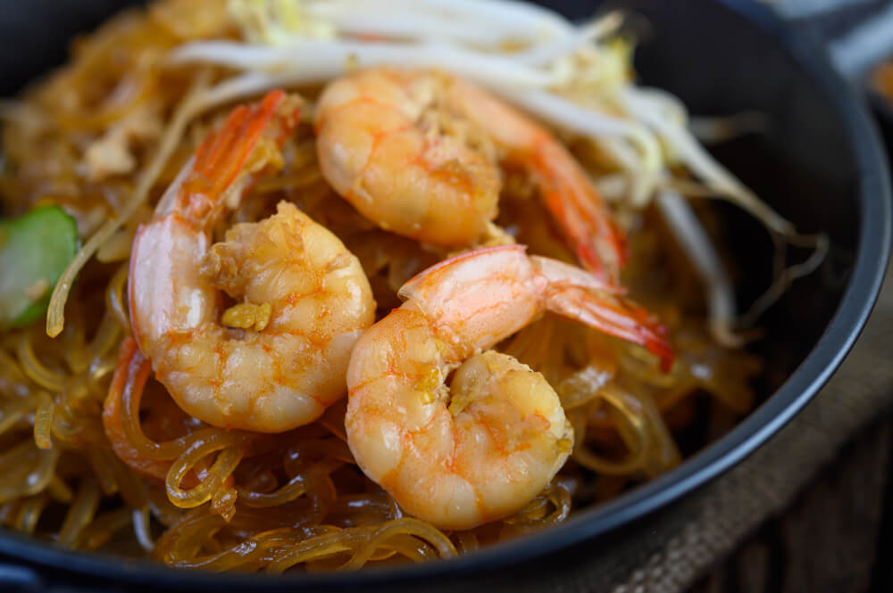 Close-up of shrimp chow mein with translucent noodles, bean sprouts, and cooked prawns in a black bowl.