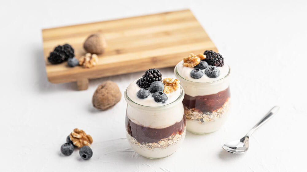 Two glass jars of chocolate overnight oats topped with yogurt, blackberries, blueberries, and walnuts, set on a white surface with a wooden cutting board and spoon nearby.