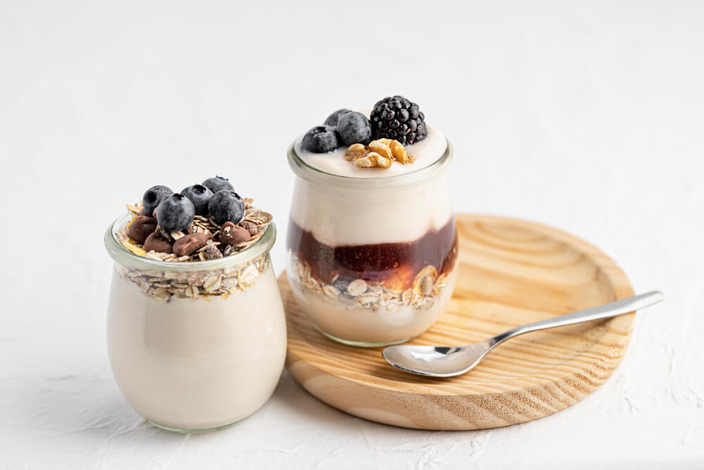 Two glass jars filled with overnight oats and yogurt, one topped with blueberries and chocolate chips, the other with berries, walnuts, and jam, placed on a wooden plate with a spoon.