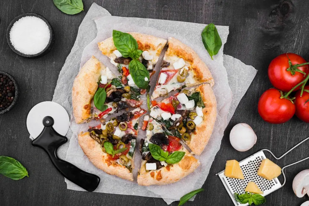 "Freshly baked veggie pizza slices on parchment paper, topped with basil, olives, spinach, mushrooms, and feta cheese, surrounded by fresh ingredients."