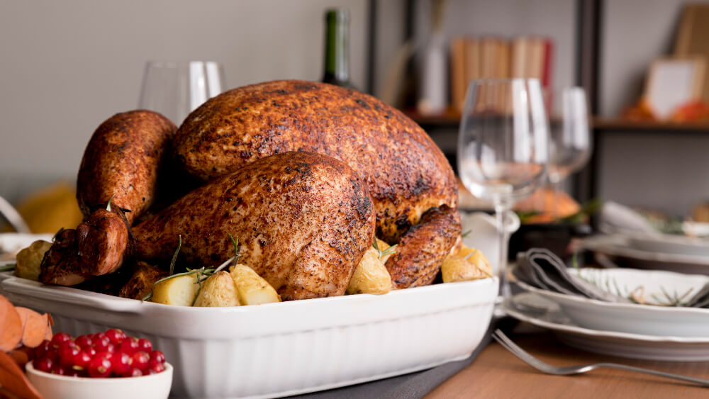 A perfectly roasted turkey served in a white ceramic dish, surrounded by golden potatoes, on an elegantly set dining table with glasses and plates.