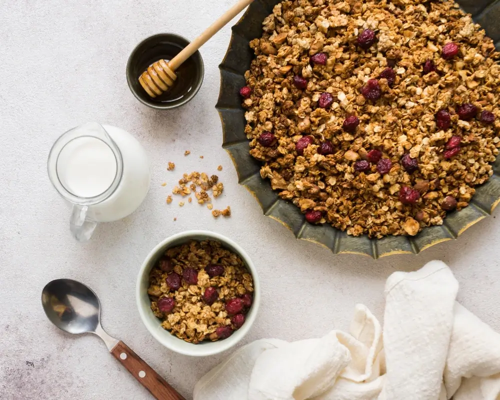 A bowl of granola with cranberries, a tray of baked granola, a jug of milk, and a honey dipper on a white surface.