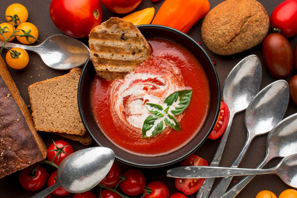 A bowl of tomato bisque soup with a swirl of cream, garnished with basil leaves and grilled bread, surrounded by fresh tomatoes, bread, and spoons.