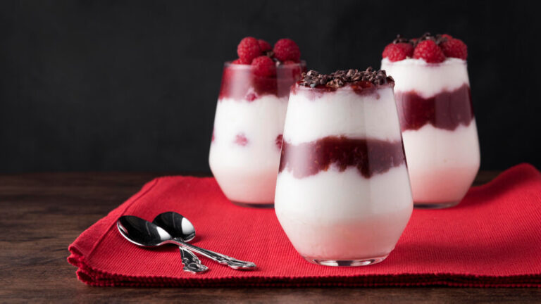 Elegant yogurt parfaits in clear glasses layered with creamy yogurt, raspberry compote, and fresh raspberries, topped with chocolate shavings, served on a red napkin with spoons.